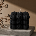 A black candle made of stacked pumpkin-shaped blocks with carved jack-o'-lantern faces, shown on a textured stone slab against a warm beige background with dried branches.