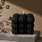 A black candle made of stacked pumpkin-shaped blocks with carved jack-o'-lantern faces, shown on a textured stone slab against a warm beige background with dried branches.