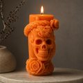An orange candle shaped like a decorative skull with floral roses, lit and standing on a stone surface against a gray wall.