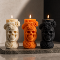 Three decorative skull-shaped pillar candles with floral carvings are lit and displayed on a stone slab against a dark background. The candles come in cream, orange, and black colors.