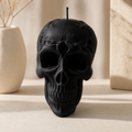 A black skull-shaped candle with a textured surface and a visible wick, styled on a light beige surface with a cream vase and stone blocks in the background. Skull Soy Wax Candle – Gothic Halloween Decor