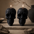 Two black skull-shaped candles with lit wicks, displayed on a stone surface in a moody, earthy setting with a ceramic vase and dried branches in the background. Skull Soy Wax Candle – Gothic Halloween Decor