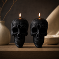 Two black skull-shaped candles with lit flames, displayed on a wooden surface with a dark, moody background and decorative vases. Skull Soy Wax Candle – Gothic Halloween Decor