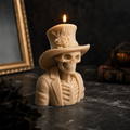 A lit cream-colored candle shaped like a skeleton wearing a top hat and suit jacket, set on a dark marble surface with gothic-themed props including a gold picture frame and old books in the background.
