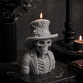 A gothic-style skeleton candle figurine wearing a top hat with a rose detail and a suit jacket, shown lit on a dark marble surface with candles and dried flowers in the background. Gray gothic skeleton candle for spooky Halloween styling