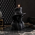 A black candle shaped like a skeleton wearing a top hat with a rose detail, lit with a small flame, displayed on a dark marble surface with gothic-themed props in the background.