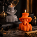 An orange Halloween-themed candle shaped like a skeleton wearing a top hat and suit jacket, lit with a flame, displayed on a dark tabletop next to a gray candle shaped like a top hat and shoulders. Orange skeleton soy wax candle for Halloween table decor