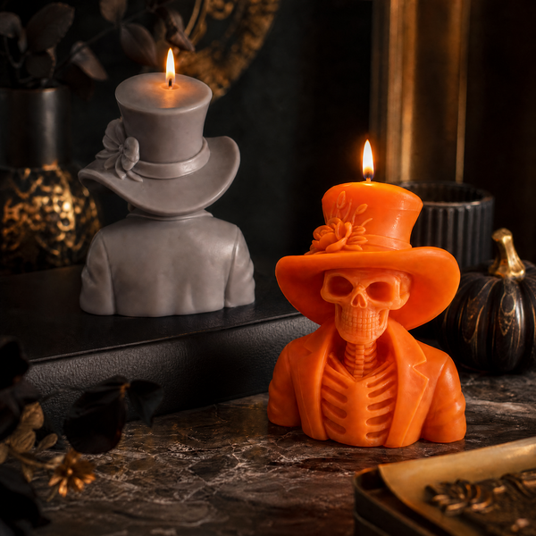 An orange Halloween-themed candle shaped like a skeleton wearing a top hat and suit jacket, lit with a flame, displayed on a dark tabletop next to a gray candle shaped like a top hat and shoulders. Orange skeleton soy wax candle for Halloween table decor
