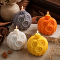 Four decorative pumpkin-shaped candles with carved sunflower details, shown lit on a stone surface with dried flowers and a vase in the background. The candles are white, gray, orange, and yellow. Fall pumpkin candles for mantel and shelf decor