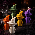 Five small witch-shaped candles with lit flames, shown in orange, white, green, yellow, and purple, arranged on a dark surface with black fabric and blurred decorative bottles in the background. Candles by Votive Candle Co