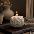 A carved pumpkin-shaped candle with a lit flame, shown on a dark wooden surface with a vase, books, and dried flowers in the background.