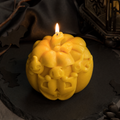 A carved yellow pumpkin candle with a lit flame, set on a dark slate surface with gothic decorative props in the background.
