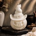A white Halloween-themed candle shaped like a pumpkin with a witch hat, displayed on a dark stone coaster with a warm, rustic background.