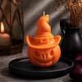 An orange Halloween candle shaped like a carved pumpkin wearing a witch hat, displayed on a black plate with a lit candle and dried flowers in the background.