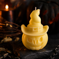 A yellow Halloween candle shaped like a carved pumpkin wearing a witch's hat, set on a dark textured surface with blurred candles and autumn decor in the background.
