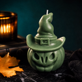 A green witch-themed candle shaped like a carved pumpkin wearing a pointed hat, styled on a dark surface with a blurred amber candle and autumn leaf in the background.
