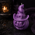 A purple Halloween-themed candle shaped like a pumpkin wearing a witch hat, photographed on a dark surface with a blurred lit lantern in the background.