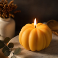 A yellow-orange pumpkin-shaped candle with a lit flame, styled on a marble surface with eucalyptus leaves and a ceramic vase in the background. Handmade Pumpkin Candle by Votive Candle Co