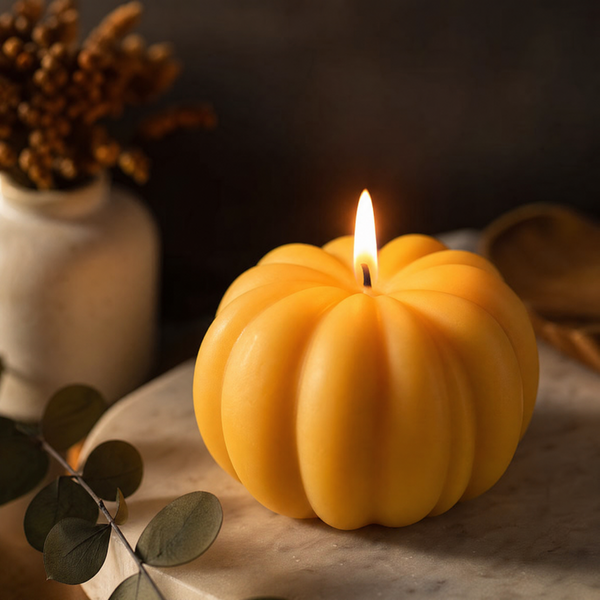 A yellow-orange pumpkin-shaped candle with a lit flame, styled on a marble surface with eucalyptus leaves and a ceramic vase in the background. Handmade Pumpkin Candle by Votive Candle Co
