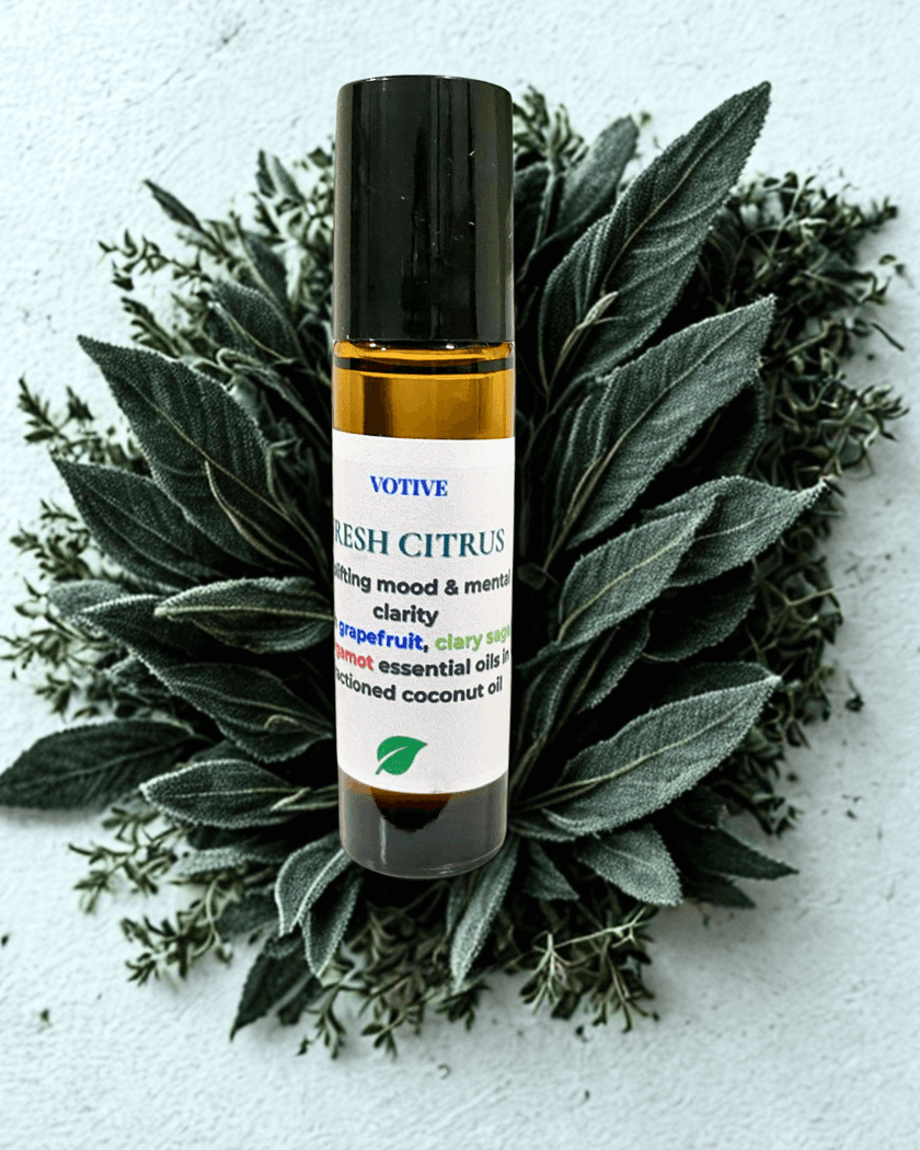 Votive Fresh Citrus essential oil roller bottle surrounded by green leaves on a light background