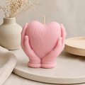 A pink heart-shaped candle cradled in two sculpted hands, with an embossed swirling pattern on the heart. The candle is styled on a round beige tray with a neutral background, a textured ceramic vase with dried flowers, and a light wood plate. Candle by Votive Candle Co