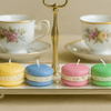 Four colorful macaron-shaped candles on a tray with floral teacups in the background.
