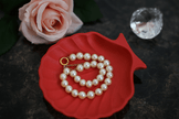 Pearl necklace on a red shell-shaped tray with a pink rose and crystal in the background.
