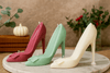 Three high-heeled shoe candles in red, green, and white on a marble surface with a neutral background.