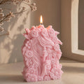 A pink sculpted flower goddess candle with a face and floral details, lit with a visible flame, set on a tabletop against a softly lit neutral background with dried flowers in the corner.
