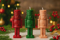 Three Christmas nutcracker-shaped candles in red, green, and peach, displayed on a tabletop with a blurred festive background of lights, pine branches, and red berries. Hand-Poured Nutcracker Candle: Sculptural Soy Wax Christmas Decor