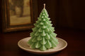 A light green Christmas tree-shaped candle with a textured, layered design sits on a small beige ceramic plate on a dark wooden table. The background is dark and features a blurred framed picture.