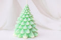 A Christmas tree-shaped candle in a light mint green color with subtle glitter details, photographed against a soft white draped fabric background.