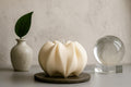 A sculptural cream-colored candle with a ribbed, pumpkin-like form sits on a dark round coaster on a tabletop. The background is styled with a small speckled ceramic vase holding a green leaf on the left and a clear glass crystal ball on a stand on the right, all against a textured light beige wall. Wavy Ribbed Candle, Soy Wax Pillar Candle, Swirl Candle by Votive Candle Co