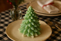 A green Christmas tree-shaped candle with layered scallop details sits on a beige ceramic plate on a black-and-white checkered tablecloth, with a blurred glass vase and plates in the background.