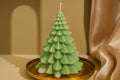 A light green Christmas tree-shaped candle with layered, scalloped leaf-like tiers and white wax at the base, displayed on a round gold tray against a warm beige background with draped fabric.
