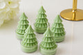 Five light green tree-shaped tea light candles in clear cups, arranged on a white surface with soft decorative flowers and a gold candlestick base in the background.