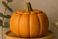 An orange pumpkin-shaped ceramic pot with a lid and a green stem, displayed on a wooden stand against a warm beige background with soft-focus greenery.