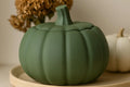 Matte green pumpkin-shaped decorative container with a lid and stem, displayed on a light wood tray with dried flowers and a small white pumpkin in the background.