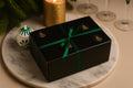 A black rectangular gift box tied with a green ribbon, placed on a round white marble tray. The scene includes a lit gold candle, pine branches, a small green-and-white ornament, and blurred wine glasses in the background. Hand-Poured Nutcracker Candle: Sculptural Soy Wax Christmas Decor