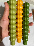Three ribbed, pumpkin-shaped candles held in a hand. Two candles are light olive green and one is bright yellow-orange, all with a hanging loop at the top.
