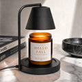 An amber glass jar candle with a white label reading 'HELLO FALL WICKLESS CANDLE' sits under a black floating lamp on a marble countertop, with books and a bowl in the background.