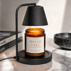 An amber glass jar candle with a white label reading 'HELLO FALL WICKLESS CANDLE' sits under a black floating lamp on a marble countertop, with books and a bowl in the background.