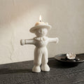 A white sculpted candle shaped like a scarecrow, lit with a small flame on top, set on a dark wooden surface against a textured beige wall. A small black dish with a white flower sits in the background. Scarecrow Soy Wax Candle – Halloween Fall Decor