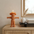 An orange terracotta scarecrow-shaped candle with a lit flame sits on a light wood console table in a styled interior setting next to a small white vase with dried grass and stacked books. Scarecrow Soy Wax Candle – Halloween Fall Decor