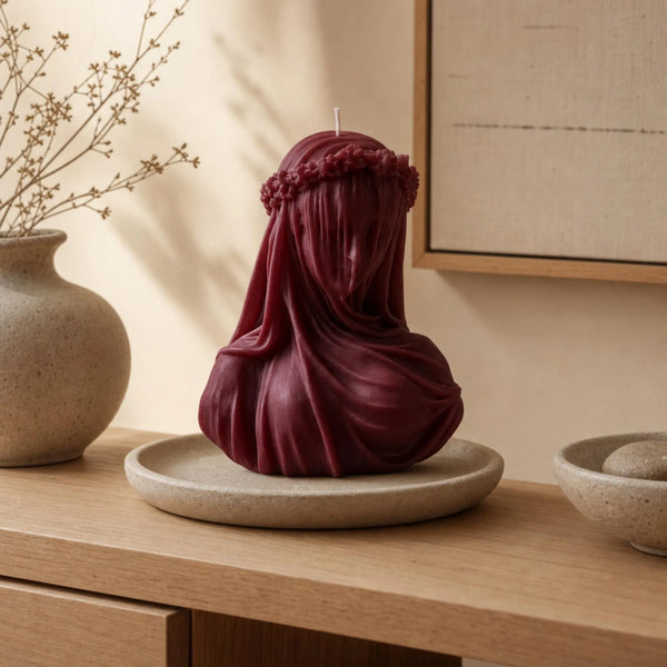 A sculptural candle shaped like a draped female figure in deep burgundy, displayed on a round beige tray on a wooden table in a warm, styled interior setting. Bordo Gothic Veiled Woman Soy Wax Candle