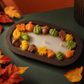 Fall Dough Bowl Candle, Handmade Soy Wax Wooden Bowl Candle An oval-shaped candle with a dark brown rim and white wax base, decorated with colorful autumn-themed wax shapes including pumpkins, acorns, leaves, and maple leaves. The candle is styled on a dark green surface with scattered red and orange autumn leaves.