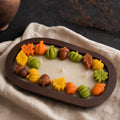 Fall Dough Bowl Candle, Handmade Soy Wax Wooden Bowl Candle An oval dark brown candle with a decorative autumn-themed wax border of pumpkins, acorns, and leaves, styled on a beige linen cloth.