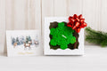 A square white gift box with a green snowflake-shaped candle inside, topped with a large red bow. A Christmas greeting card with reindeer and the words 'Merry Christmas' stands beside it on a light surface against a pale wood plank background, with a green pine branch at the right edge. Snowflake Candle Gift Set, Handmade Soy Wax Christmas Candles, Winter Decor