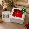 A red snowflake-shaped candle in a white gift box with a green bow, shown beside a Christmas greeting card on a tabletop with holiday decor in the background. Snowflake Candle Gift Set, Handmade Soy Wax Christmas Candles, Winter Decor