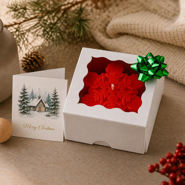 A red snowflake-shaped candle in a white gift box with a green bow, shown beside a Christmas greeting card on a tabletop with holiday decor in the background. Snowflake Candle Gift Set, Handmade Soy Wax Christmas Candles, Winter Decor